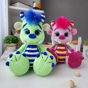 May include: Two handmade crochet monster dolls. One is green with blue accents and the other is pink with yellow accents. Both have large eyes, horns, and fluffy hair. The green monster has blue and white striped details.