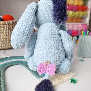 May include: A light blue plush donkey toy with a dark blue mane and tail. The donkey has a pink bow and a button on its back. The toy is handmade and has a soft, textured appearance.