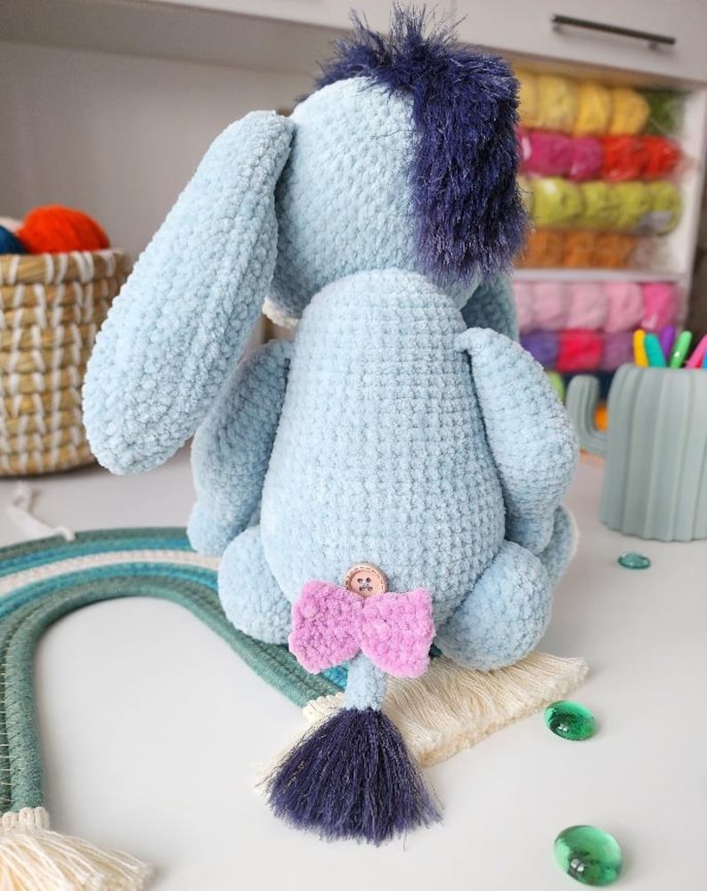 May include: A light blue plush donkey toy with a dark blue mane and tail. The donkey has a pink bow and a button on its back. The toy is handmade and has a soft, textured appearance.