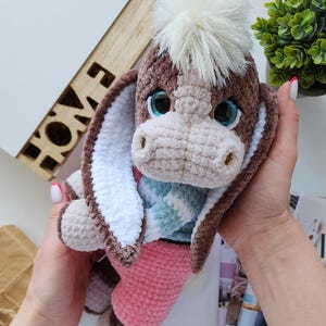 May include: A handmade crochet donkey plush toy. The donkey has brown and beige fur, large blue eyes, and a fluffy white mane. It wears a pink bottom and a blue and white patterned scarf. The toy is held in a person's hands.