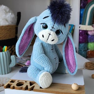 May include: A handmade plush toy of Eeyore, the donkey from Winnie the Pooh. The blue donkey has large, expressive eyes, a dark blue hair tuft, and pink-lined ears. The plush is sitting on a wooden surface.