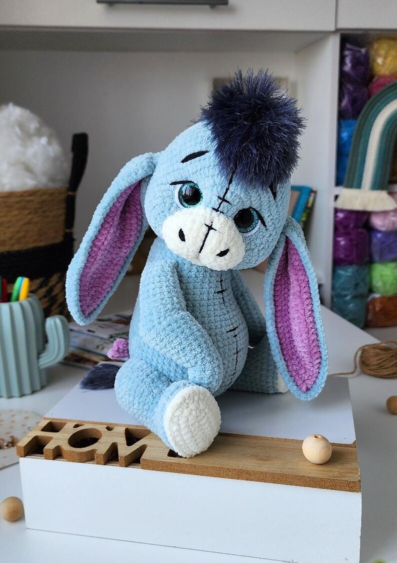 May include: A handmade plush toy of Eeyore, the donkey from Winnie the Pooh. The blue donkey has large, expressive eyes, a dark blue hair tuft, and pink-lined ears. The plush is sitting on a wooden surface.