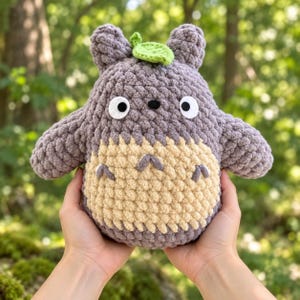May include: A grey and beige crocheted plush toy featuring a rounded body, two pointed ears, a small green leaf on top, and a cream-coloured belly with three dark grey chevron markings. The face has two white circular eyes with black pupils.