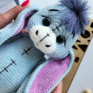 May include: A handmade plush toy in the shape of a donkey, featuring a light blue body, purple inner ears, and a fluffy blue pom-pom. The toy has stitched details and large, glittery eyes. A charming, soft toy.