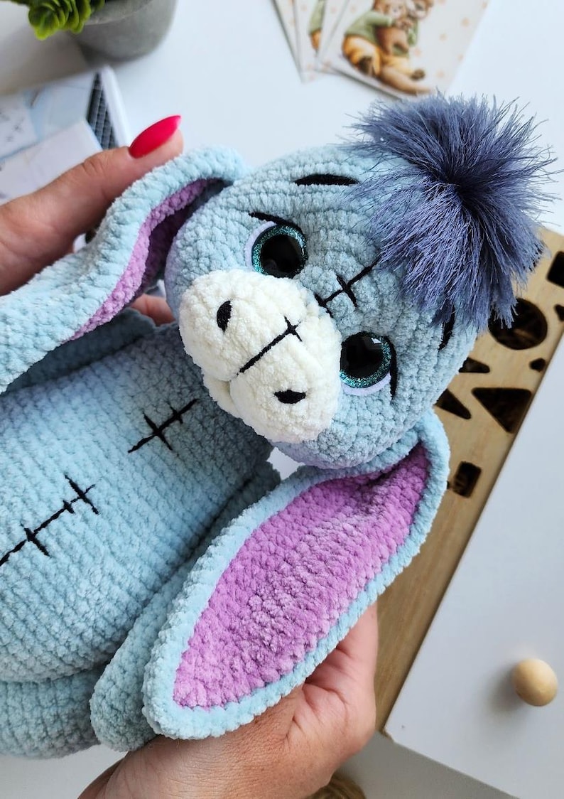 May include: A handmade plush toy in the shape of a donkey, featuring a light blue body, purple inner ears, and a fluffy blue pom-pom. The toy has stitched details and large, glittery eyes. A charming, soft toy.