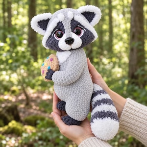 May include: A crocheted raccoon plush toy with gray and white fur, large dark eyes, and a black nose. The raccoon has a striped tail and is holding a small, round, tan-colored cookie decorated with colorful sprinkles in its dark gray paws.