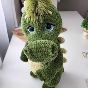 Puede incluir: Un peluche de dinosaurio verde hecho a mano con ganchillo, con una cresta verde claro y ojos azules. El peluche tiene un aspecto texturizado y suave y una barriga de color crema. El dinosaurio está de pie, mostrando sus brazos y piernas.