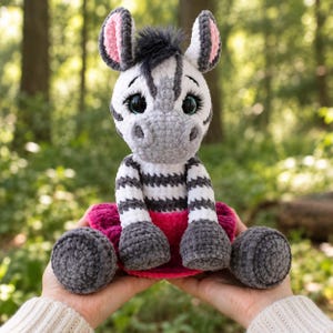 May include: A crocheted zebra plush toy featuring a white and gray striped body, a dark gray mane, large black eyes with long eyelashes, pink inner ears, and a bright magenta skirt. The stuffed animal is crafted from soft yarn with a textured finish.
