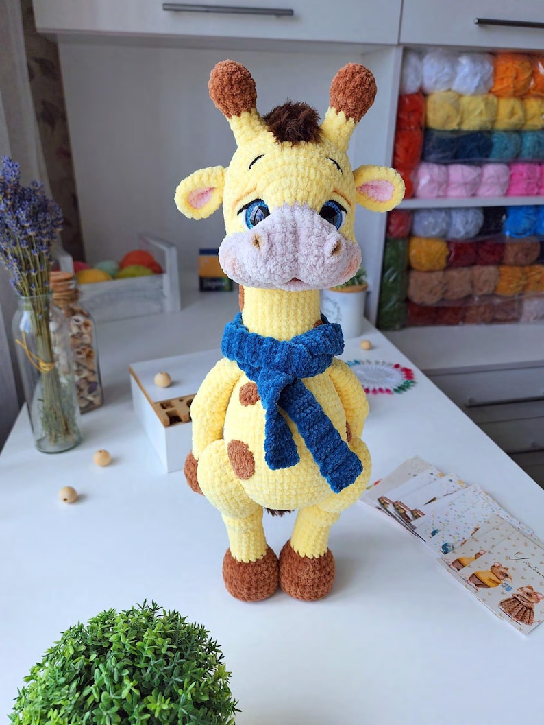 May include: A handmade crochet giraffe plush toy with yellow yarn, brown spots, and a textured blue scarf. The giraffe features large blue eyes, a light pink muzzle, and brown horn tips. The soft, plush construction gives it a rounded, friendly appearance.