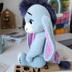 May include: A light blue plush donkey toy with a dark blue mane and tail, and pink inner ears. The toy is sitting on a wooden base that says "HOME". The donkey has a sad expression and is made of soft, textured material.