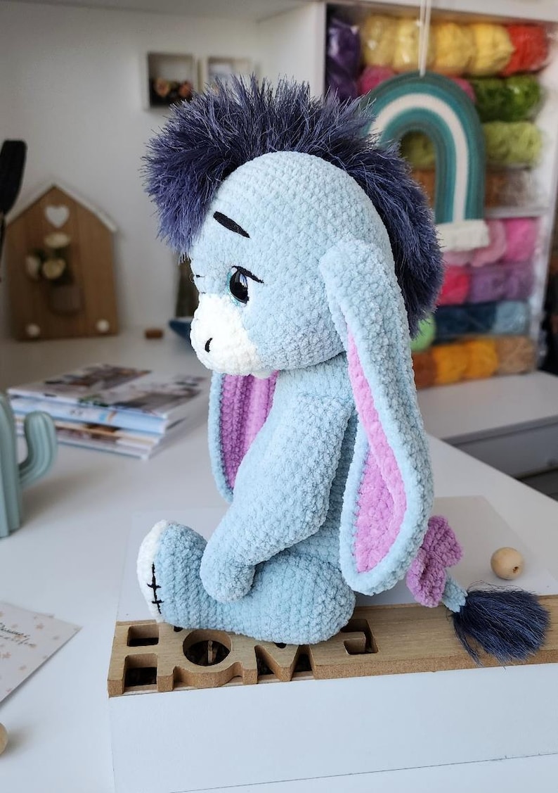 May include: A light blue plush donkey toy with a dark blue mane and tail, and pink inner ears. The toy is sitting on a wooden base that says "HOME". The donkey has a sad expression and is made of soft, textured material.