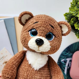 May include: A handmade brown teddy bear with a fluffy texture. The bear has large, expressive blue eyes with black pupils, a black nose, and a white chest. The ears are brown with white inner details. A charming, cuddly toy.