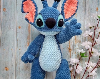 May include: Blue and light blue crocheted plush toy with large ears, a black nose, and big eyes. The toy is standing on a wooden surface with white flowers.