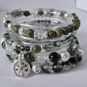 Könnte beinhalten: Mehrere Armbänder mit grünen, transparenten und silbernen Perlen. Ein Armband hat einen silbernen Kompass-Anhänger. Die Armbänder werden auf einem transparenten Glasbehälter präsentiert.