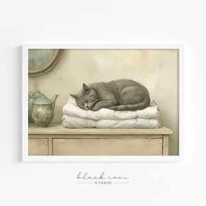 May include: A framed print featuring a sleeping gray cat atop a stack of white blankets. The cat is curled up, eyes closed, in a relaxed pose. The print is set against a neutral background with a vintage aesthetic, with a teapot and mirror visible.