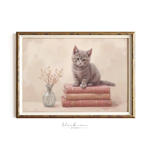 May include: A framed print featuring a gray kitten sitting atop a stack of three pink books. A clear glass vase with dried flowers is to the left. The frame is gold-toned, and the background is a soft beige.