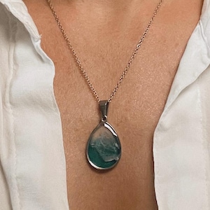 May include: A silver-toned pendant necklace featuring a teardrop-shaped pendant. The pendant has a teal and white abstract design encased in a silver frame. The necklace is worn against a white shirt.