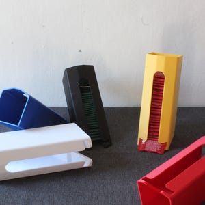 Op de afbeelding: Vijf verschillende gekleurde plastic containers voor het bewaren van theezakjes. De containers zijn in verschillende vormen en maten. De containers staan op een grijs oppervlak.