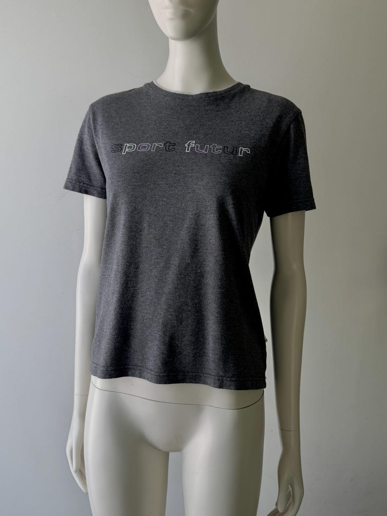 Peut inclure: T-shirt gris fonc&eacute; avec l'inscription "sport futur" en blanc soulign&eacute; sur la poitrine. Le t-shirt a des manches courtes et un col rond. Le v&ecirc;tement est pr&eacute;sent&eacute; sur un mannequin.