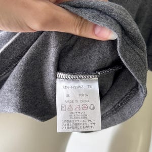 Peut inclure: Gros plan sur un t-shirt gris avec une &eacute;tiquette. L'&eacute;tiquette indique "ATN-4435WZ TE", "綿 100%" et "MADE IN CHINA". L'&eacute;tiquette comprend &eacute;galement des instructions de lavage et du texte japonais. Le t-shirt est fait d'un tissu doux et textur&eacute;.