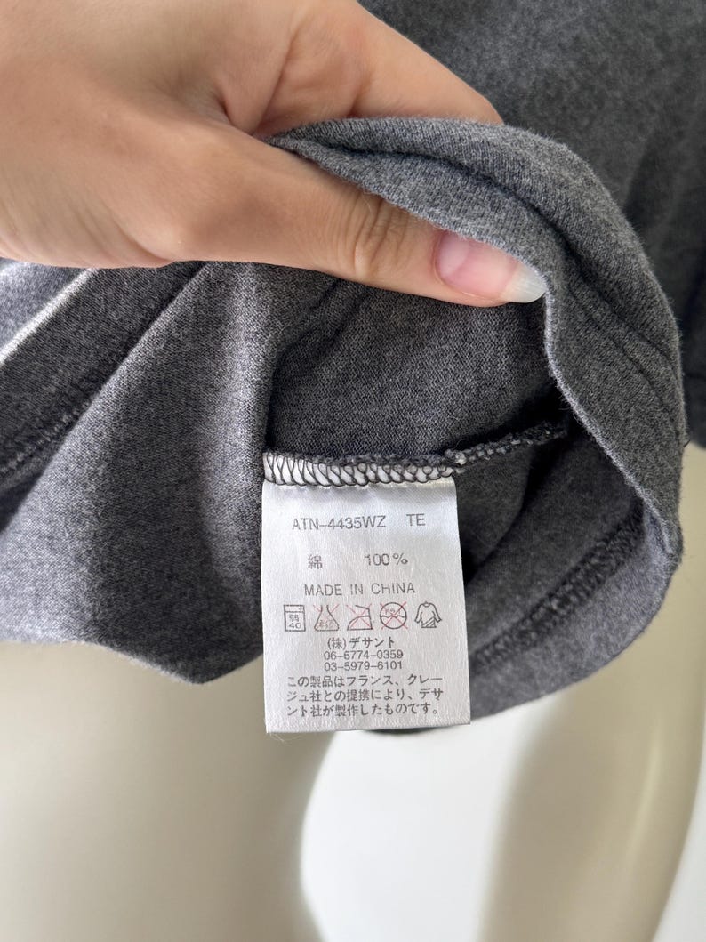 Peut inclure: Gros plan sur un t-shirt gris avec une &eacute;tiquette. L'&eacute;tiquette indique "ATN-4435WZ TE", "綿 100%" et "MADE IN CHINA". L'&eacute;tiquette comprend &eacute;galement des instructions de lavage et du texte japonais. Le t-shirt est fait d'un tissu doux et textur&eacute;.