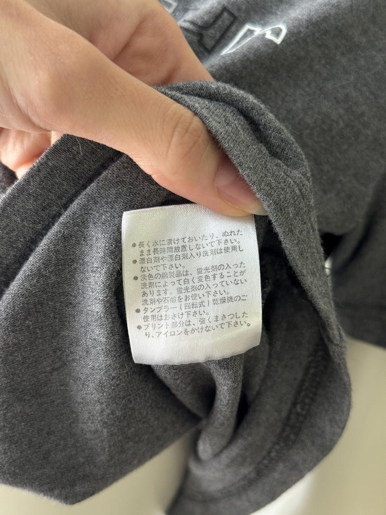 Peut inclure: Gros plan sur un t-shirt gris fonc&eacute; avec une &eacute;tiquette blanche. L'&eacute;tiquette contient du texte japonais avec des instructions d'entretien. Le t-shirt est fait d'un tissu doux et textur&eacute;.