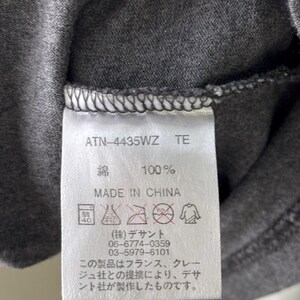 Peut inclure: Gros plan sur une &eacute;tiquette de v&ecirc;tement gris fonc&eacute; avec du texte et des symboles d'entretien. L'&eacute;tiquette indique "ATN-4435WZ TE", "綿 100%" et "MADE IN CHINA". Du texte japonais et des instructions d'entretien sont &eacute;galement visibles.