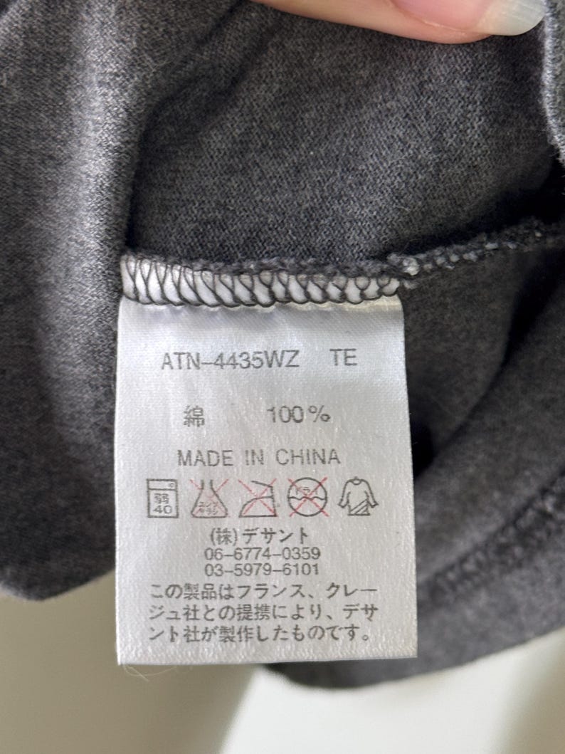 Peut inclure: Gros plan sur une &eacute;tiquette de v&ecirc;tement gris fonc&eacute; avec du texte et des symboles d'entretien. L'&eacute;tiquette indique "ATN-4435WZ TE", "綿 100%" et "MADE IN CHINA". Du texte japonais et des instructions d'entretien sont &eacute;galement visibles.