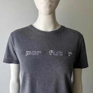 Peut inclure: Un t-shirt gris avec l'inscription "sport futur" en blanc et violet. Le t-shirt a des manches courtes et une encolure blanche. Le t-shirt est pr&eacute;sent&eacute; sur un mannequin.