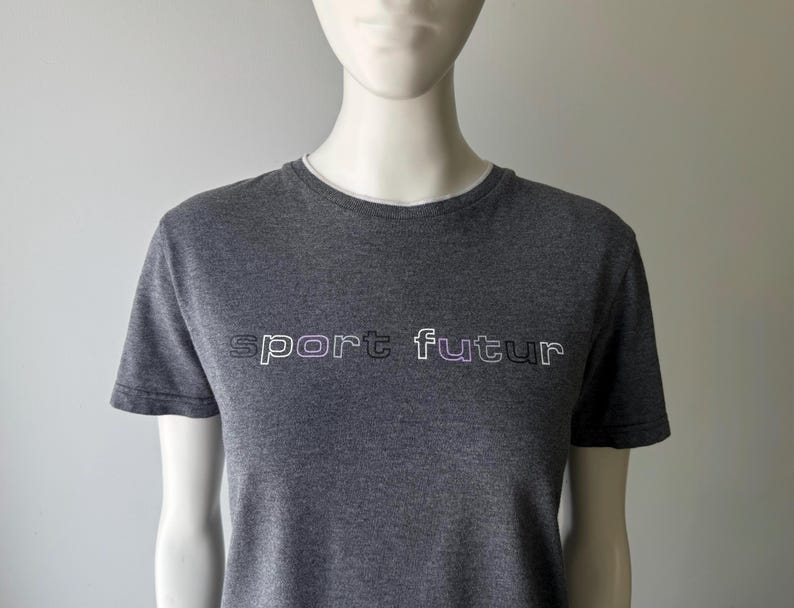 Peut inclure: Un t-shirt gris avec l'inscription "sport futur" en blanc et violet. Le t-shirt a des manches courtes et une encolure blanche. Le t-shirt est pr&eacute;sent&eacute; sur un mannequin.