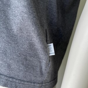 Peut inclure: Gros plan sur un t-shirt gris. Le tissu est gris chin&eacute;, avec une texture subtile. Une petite &eacute;tiquette rectangulaire est cousue dans la couture lat&eacute;rale. L'&eacute;tiquette a un fond blanc avec un logo.