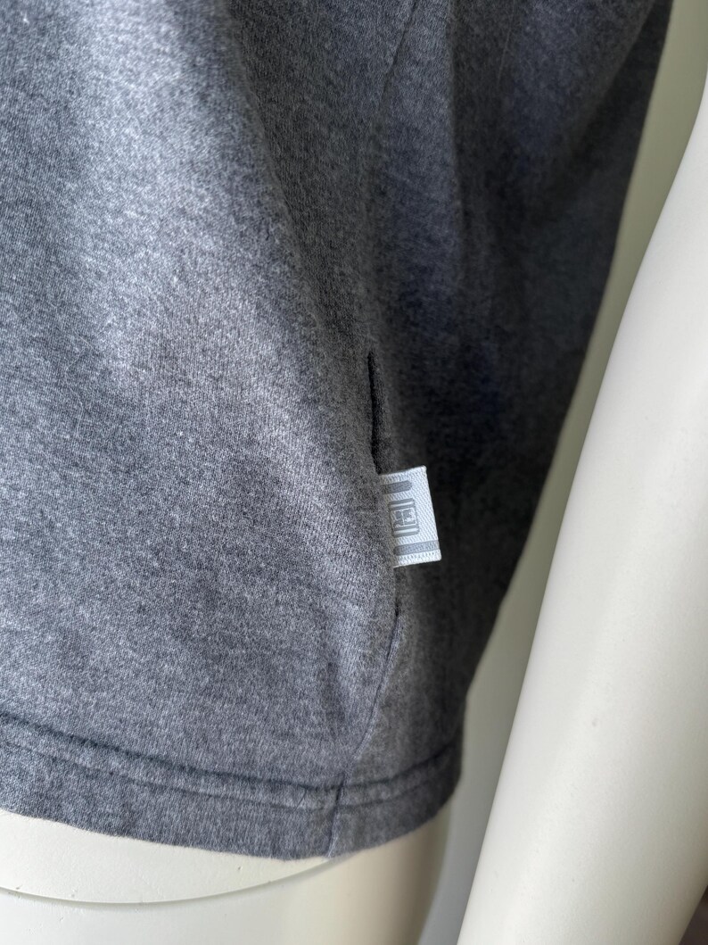 Peut inclure: Gros plan sur un t-shirt gris. Le tissu est gris chin&eacute;, avec une texture subtile. Une petite &eacute;tiquette rectangulaire est cousue dans la couture lat&eacute;rale. L'&eacute;tiquette a un fond blanc avec un logo.