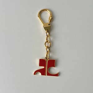 Op de afbeelding: Een gouden sleutelhanger met een rode emaille hanger met de letters "AC" in een gestileerd lettertype.