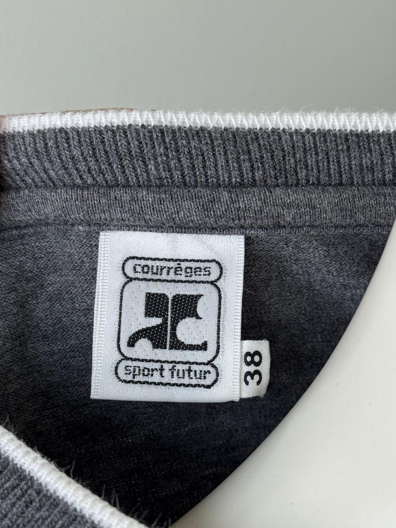 Peut inclure: Gros plan sur un col de sweat-shirt gris avec une bordure blanche. Une &eacute;tiquette blanche affiche le logo "Courr&egrave;ges" et le texte "sport futur". Le num&eacute;ro "38" est imprim&eacute; sur l'&eacute;tiquette.