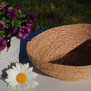 May include: A woven basket with a natural, light brown color sits next to a gray and white patterned planter filled with purple flowers. A single white daisy with a yellow center rests nearby. The scene is set outdoors.