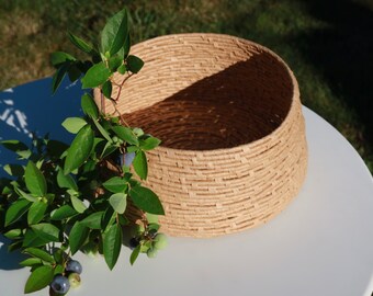 May include: A round, light tan woven basket with a textured, horizontal coil pattern. A leafy green branch with small, round, dark blue and light green berries is draped over the side of the basket, resting against its exterior wall.