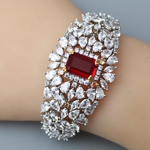 May include: A silver-toned bracelet with a large, rectangular, deep red gemstone at its center. The gemstone is surrounded by smaller, clear, teardrop-shaped stones. The bracelet is a statement piece, likely for special occasions.