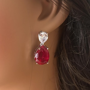 May include: A pair of silver-toned drop earrings. Each earring features a pear-shaped, deep red gemstone suspended below a smaller, clear, teardrop-shaped stone. The earrings are set against a blurred background.