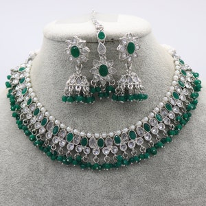 May include: A silver necklace and earrings set with green and clear stones. The necklace has a layered design with green beads and white pearls. The earrings are dangle style with a green stone and clear stones.