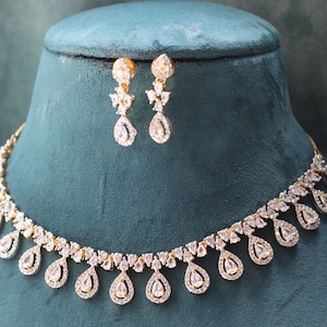 May include: A gold-toned necklace and matching earrings set. The jewelry features a series of teardrop-shaped, clear gemstones. The necklace and earrings are displayed on a teal-colored bust, showcasing the intricate design and sparkle.