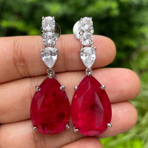 May include: A pair of silver-toned drop earrings featuring a cluster of clear, round-cut stones at the top, a pear-shaped clear stone in the middle, and a large, teardrop-shaped red stone at the bottom. The earrings are displayed against a green background.