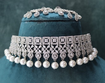 May include: A silver-toned bridal jewelry set featuring a choker necklace with square and floral designs, accented with dangling faux pearls. The set includes a matching tiara, all displayed on a teal velvet surface.