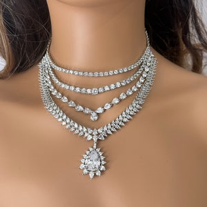 May include: A silver multi-layered necklace with a large pear-shaped pendant. The necklace is made of cubic zirconia stones and has a delicate, elegant design.