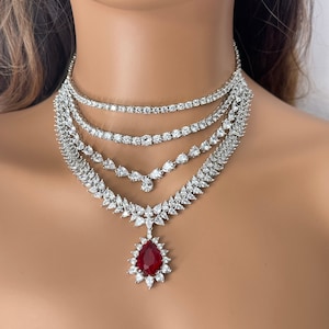 Könnte beinhalten: Mehrschichtige silberne Halskette mit klaren und roten Edelsteinen. Die Halskette hat mehrere Stränge aus funkelnden, klaren Steinen, mit einem großen, tropfenförmigen roten Edelsteinanhänger in der Mitte.