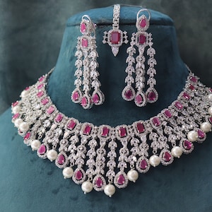Puede incluir: Un collar y pendientes de plata con piedras rosas y perlas blancas. El collar tiene un diseño en capas con una fila central de piedras rosas más grandes y piedras blancas más pequeñas.