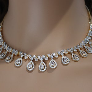 May include: A gold-toned necklace with a series of clear, teardrop-shaped pendants. Each pendant is surrounded by smaller, clear stones, creating a sparkling effect. The necklace is displayed on a neutral background.