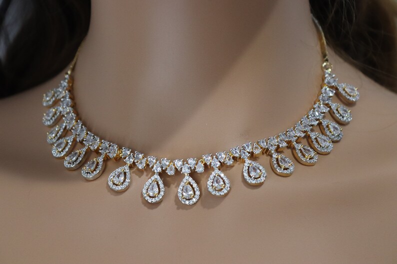 May include: A gold-toned necklace with a series of clear, teardrop-shaped pendants. Each pendant is surrounded by smaller, clear stones, creating a sparkling effect. The necklace is displayed on a neutral background.