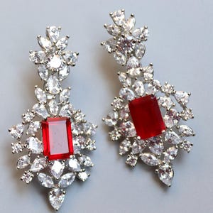 Peut inclure: Une paire de boucles d'oreilles argentées avec un motif floral. Chaque boucle d'oreille présente une grande pierre rouge rectangulaire au centre, entourée de nombreuses pierres claires et scintillantes. Les boucles d'oreilles sont pour oreilles percées.
