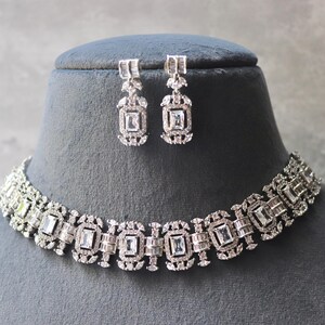 May include: A silver-toned necklace and matching earrings set. The jewelry features a repeating pattern of rectangular and square-shaped clear stones. The necklace and earrings are displayed on a black mannequin head against a gray background.