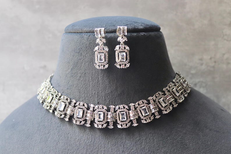 May include: A silver-toned necklace and matching earrings set. The jewelry features a repeating pattern of rectangular and square-shaped clear stones. The necklace and earrings are displayed on a black mannequin head against a gray background.